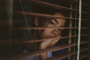 A sad looking young woman looking out a window through the open blinds.