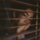 A sad looking young woman looking out a window through the open blinds.