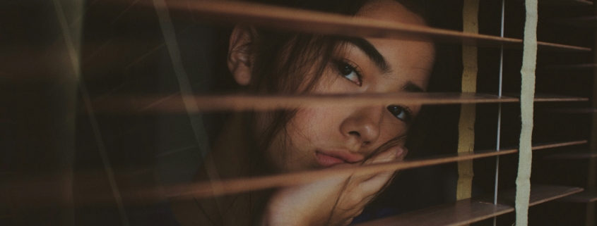 A sad looking young woman looking out a window through the open blinds.