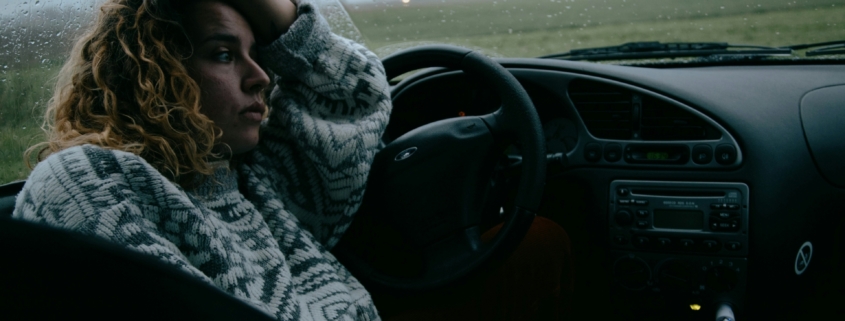 A woman sitting in a parked car with rain on the windshield.