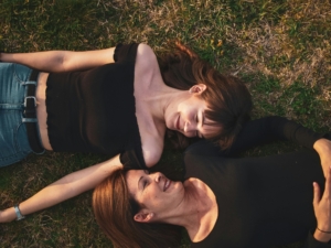 two women laying on grass outside on their backs, with their heads turned looking at each other.