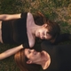 two women laying on grass outside on their backs, with their heads turned looking at each other.