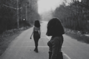 Two women with similar hair and outfits standing in the middle of a wooded road, in black and white.