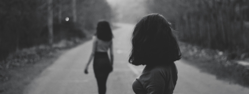 Two women with similar hair and outfits standing in the middle of a wooded road, in black and white.