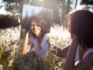 A woman in a field, looking at her reflection in a mirror.