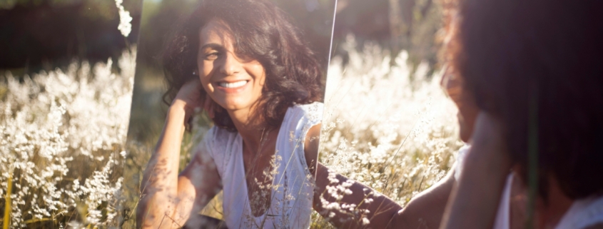 A woman in a field, looking at her reflection in a mirror.
