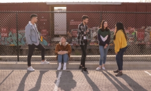 A group of young people standing outside in front of a fence by a railyard.