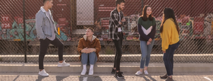 A group of young people standing outside in front of a fence by a railyard.