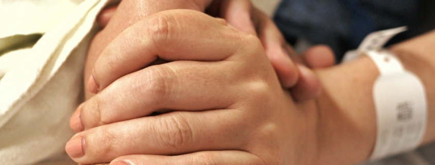 A close up of two white people holding hands. One person is wearing a hospital bracelet.