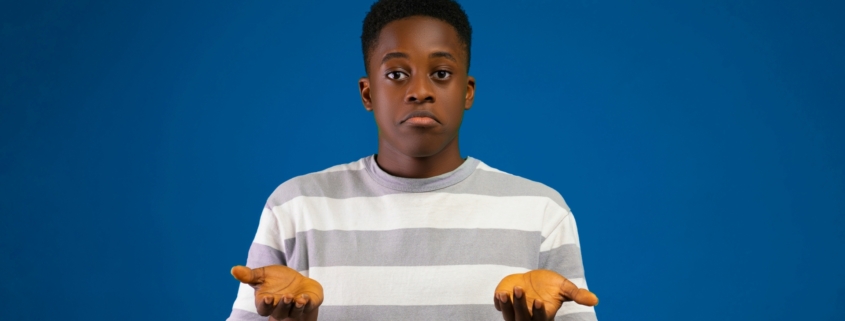 A Black teenage boy, shrugging his shoulders with his hands in the air like he's confused.