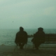 Two people sitting on a bench in front of a body of water, from behind.