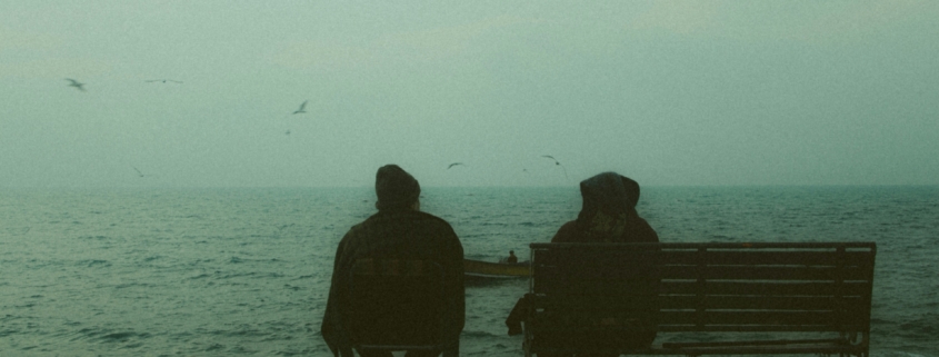 Two people sitting on a bench in front of a body of water, from behind.