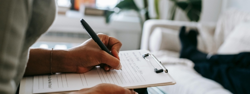 A therapist taking notes on a clipboard, across from a person laying on a couch, who we can only see from the legs down.