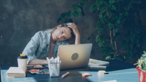 A woman looking distressed with her head in her hand, looking at her computer, sitting at a desk surrounded by office supplies.