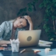 A woman looking distressed with her head in her hand, looking at her computer, sitting at a desk surrounded by office supplies.