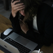 A light skinned woman sitting at a computer in a dark room with her head in her hands like she's upset.