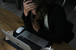A light skinned woman sitting at a computer in a dark room with her head in her hands like she's upset.