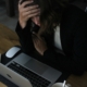 A light skinned woman sitting at a computer in a dark room with her head in her hands like she's upset.