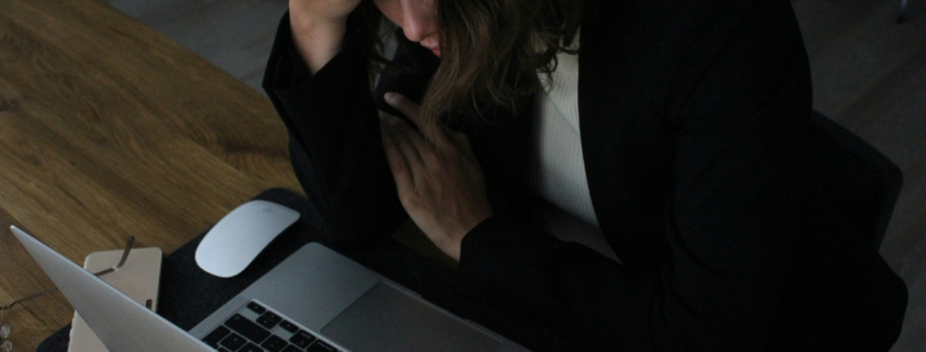 A light skinned woman sitting at a computer in a dark room with her head in her hands like she's upset.