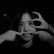 A black and white photo of a woman whose face is being pulled into a smile by hands from outside of the frame.