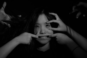 A black and white photo of a woman whose face is being pulled into a smile by hands from outside of the frame.
