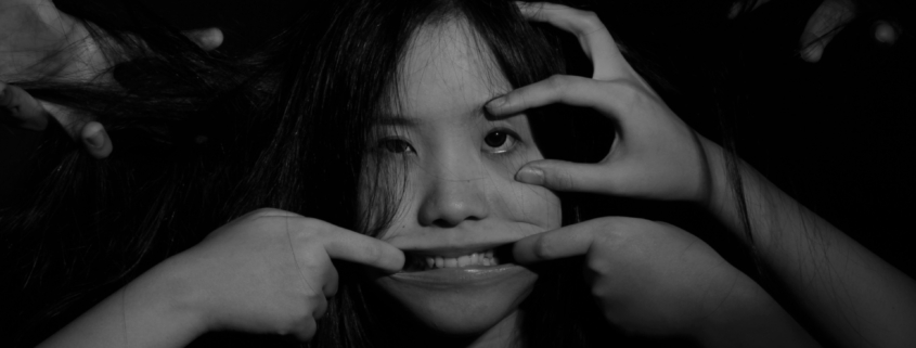 A black and white photo of a woman whose face is being pulled into a smile by hands from outside of the frame.