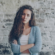 A white woman with long curly hair standing in front of a brick wall with her arms crossed, with her lips pursed.
