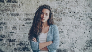 A white woman with long curly hair standing in front of a brick wall with her arms crossed, with her lips pursed.