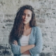A white woman with long curly hair standing in front of a brick wall with her arms crossed, with her lips pursed.