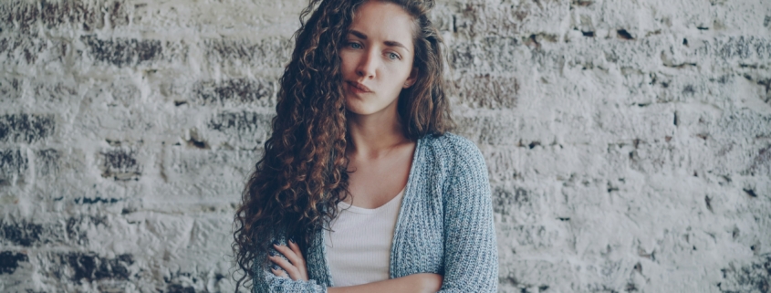 A white woman with long curly hair standing in front of a brick wall with her arms crossed, with her lips pursed.