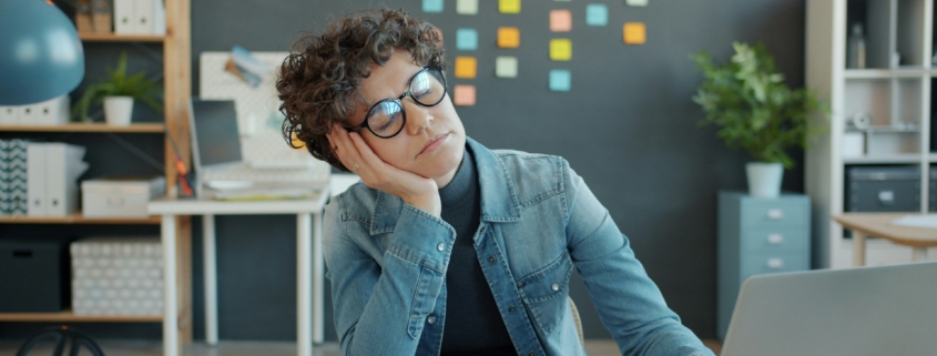 A tired person with light skin and short curly brown hair, leaning their head on their hand at their desk, with a notebook and laptop open in front of them. On the wall behind them is a bookcase and post-it notes on the wall.