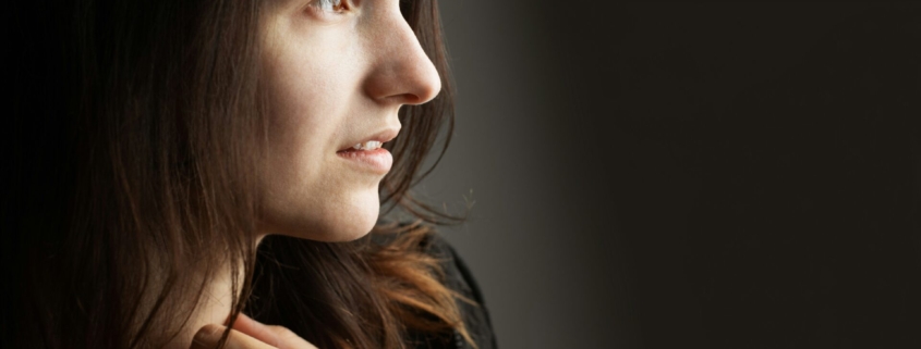 A woman with light skin in a black sweater standing in front of a dark wall, holding her hand in front of her chest.