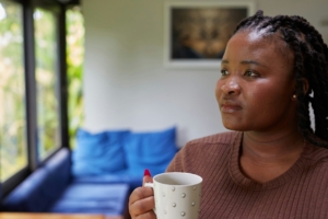 A young Black woman standing in a living room in front of the windows with a mug of coffee, looking thoughtful.