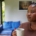 A young Black woman standing in a living room in front of the windows with a mug of coffee, looking thoughtful.