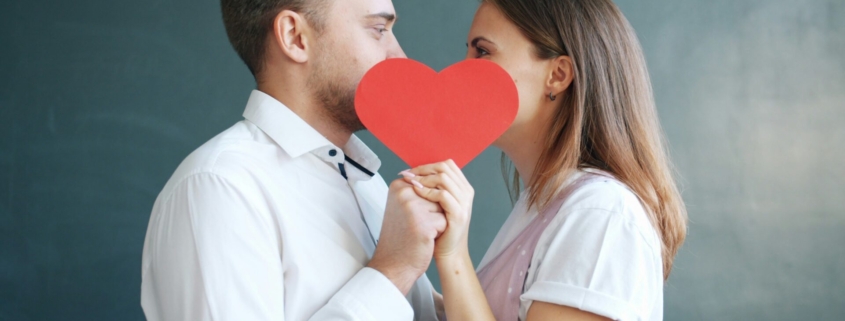 A white man and woman standing in front of a green wall facing each other, holding a red paper heart in front of their faces.