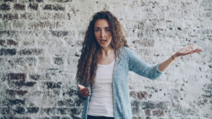 A light skinned woman with dark blonde curly hair standing in front of a brick wall, holding her arms up like she's frustrated.