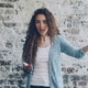 A light skinned woman with dark blonde curly hair standing in front of a brick wall, holding her arms up like she's frustrated.