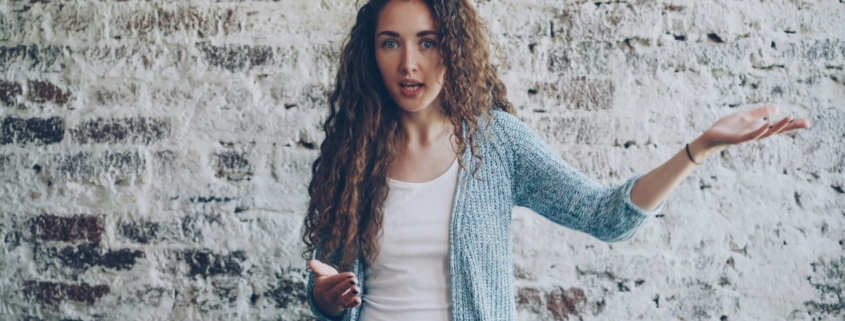 A light skinned woman with dark blonde curly hair standing in front of a brick wall, holding her arms up like she's frustrated.