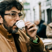 A middle aged or older man with dark brown hair and a light brown jacket drinking out of a white espresso cup outdoors in a city.