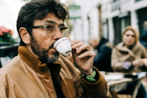 A middle aged or older man with dark brown hair and a light brown jacket drinking out of a white espresso cup outdoors in a city.