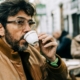 A middle aged or older man with dark brown hair and a light brown jacket drinking out of a white espresso cup outdoors in a city.