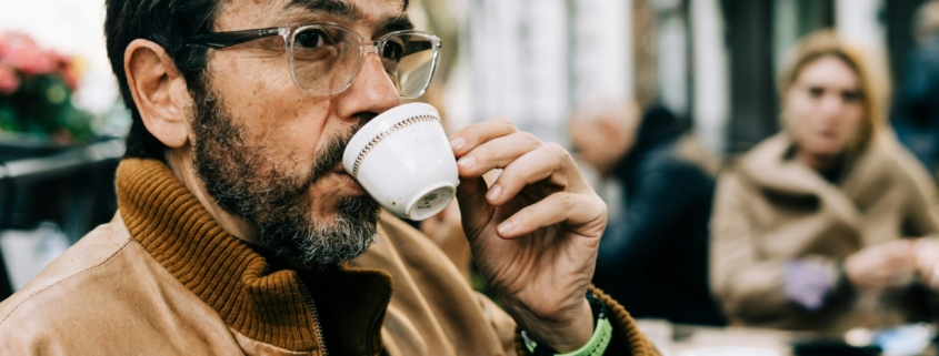 A middle aged or older man with dark brown hair and a light brown jacket drinking out of a white espresso cup outdoors in a city.