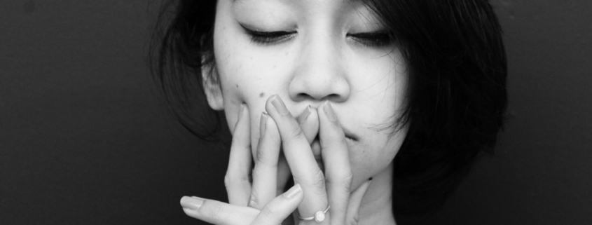 A black and white photo of an Asian woman looking sad and holding her hands up to her mouth like she's thinking.