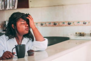 A woman with brown skin and black hair sitting at a counter with a cup of coffee and a muffin in front of her, with her head in her hand and her eyes closed, looking frustrated.