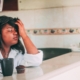 A woman with brown skin and black hair sitting at a counter with a cup of coffee and a muffin in front of her, with her head in her hand and her eyes closed, looking frustrated.