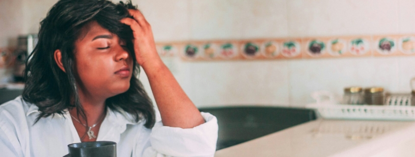 A woman with brown skin and black hair sitting at a counter with a cup of coffee and a muffin in front of her, with her head in her hand and her eyes closed, looking frustrated.