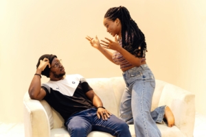A Black man and woman sitting on a couch in front of a light yellow background, looking like they're arguing.