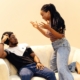 A Black man and woman sitting on a couch in front of a light yellow background, looking like they're arguing.