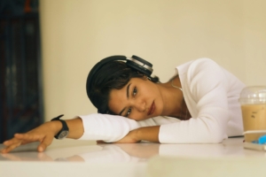 A young South Asian woman with headphones on laying her torso on a table indoors, looking distressed. 