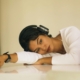 A young South Asian woman with headphones on laying her torso on a table indoors, looking distressed.